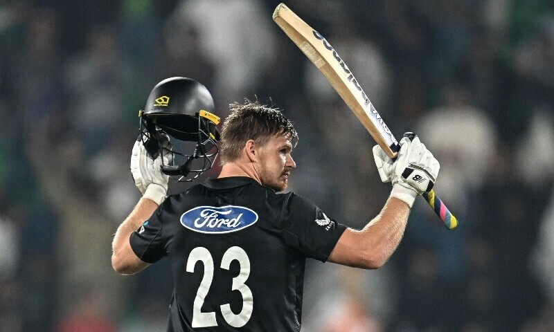 A cricketer wearing a black jersey with the number 23 holds up a bat and helmet while celebrating in front of a cheering crowd.