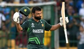 A cricketer wearing a green jersey raises his bat and helmet in triumph, surrounded by a cheering crowd.